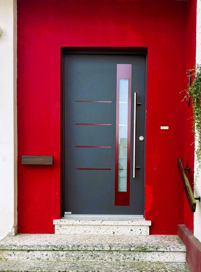 Austausch Haustür Graue Haustür mit Glaselementen vor roter Wand und Treppe.